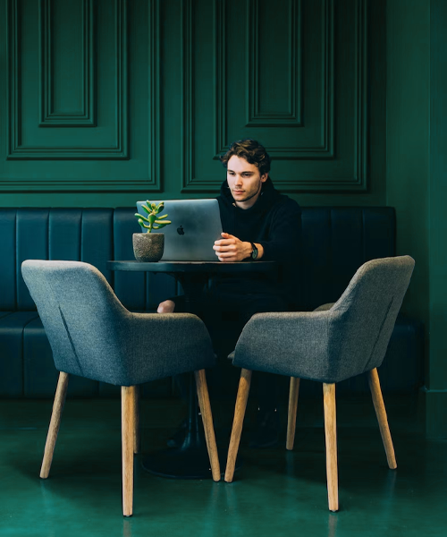 Ung man arbetar på en bärbar dator vid ett cafébord med en liten krukväxt, omgiven av två grå stolar mot en grön vägg